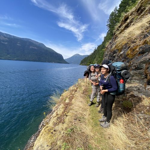 Participants enjoy the view as they stand at the edge of a vast, blue lake, which is bordered by tall cliffs and hills