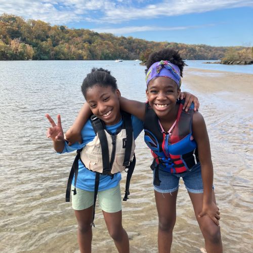 Two young participants pose for the camera with large smiles and arms around each other. They are wearing life jackets and standing in the water during a Mountaineer Canoe trip in DC.