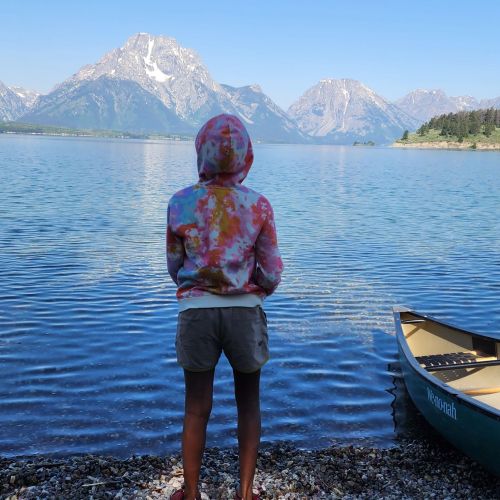 A participant has their back facing the camera as they gaze upon a clear, blue lake in front of them. An empty canoe rests on the shore beside them and mountains are in the distance.
