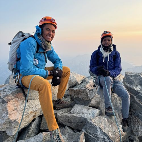 Two participants in climbing gear sit on top of stacks of large rocks to celebrate a successful climb in Grand Teton, Wyoming.