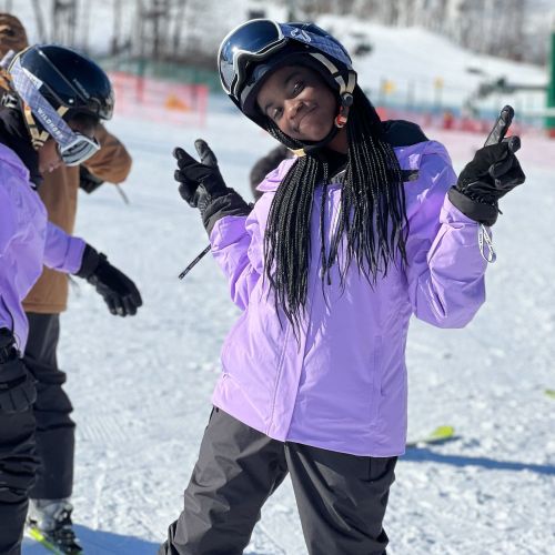 A participant in a bright purple jacket and winter gear poses with peace signs and a bright smile, showing her enjoyment during a Snow Club event on a ski hill.