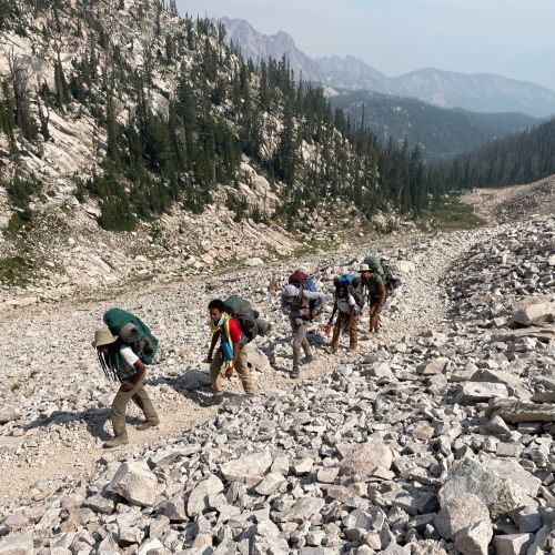 Five hikers follow a rocky incline during a backpacking trip in the Sawtooth Mountains. The trail itself is long and extends far into the background.