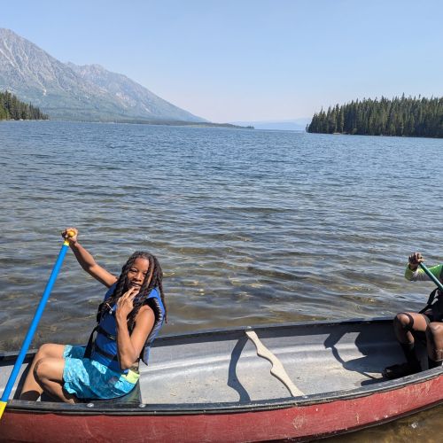 Two participants work together as they canoe in Jackson Lake, Wyoming. The vast, clear lake extends to the far background, with lines of trees and mountains in the distance.