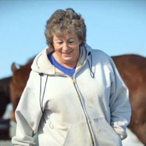 A woman smiles as she looks down; surrounding her are brown horses and a clear blue sky.