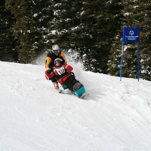 During adaptive racing, a CDA staff member guides a participant wearing a racing bib number as they slide down a snowy hill in a bi-ski.