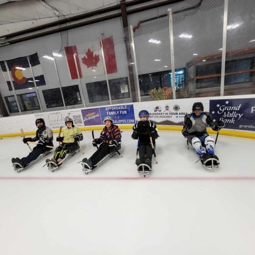 Five participants form a team and gear up for a sled hockey scrimmage. They are seated on their sleds on the ice inside an arena.