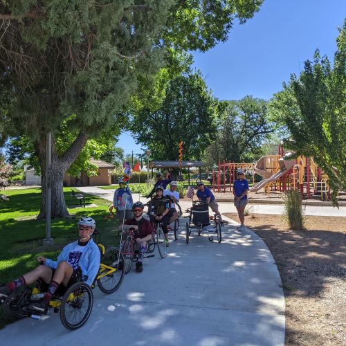  A group of participants cycle with trikes along the Colorado River front trail, which includes a shaded paved path for easy biking. They are biking past a playground under a canopy of trees.