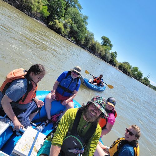 Participants wearing life jackets enjoy river rafting during a clear sunny day. The river itself is vast and lined with dense, green trees.