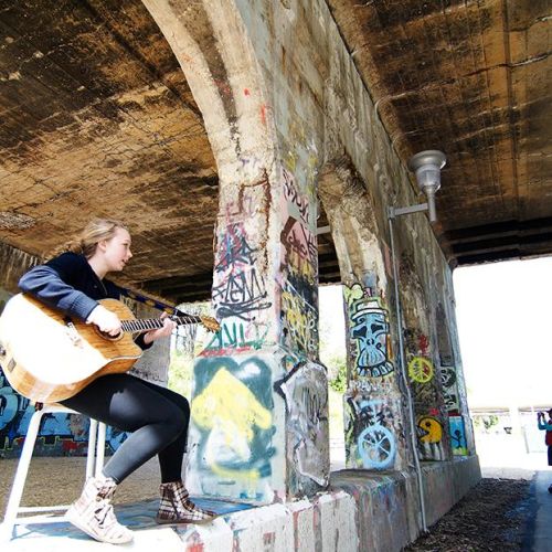 A young adult is sitting and playing guitar beneath a graffitied overpass.