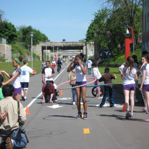 Under a sunny sky, a group of children and teens gathered on the path are engaged in outdoor recreational activities like hula hooping and jumping rope.