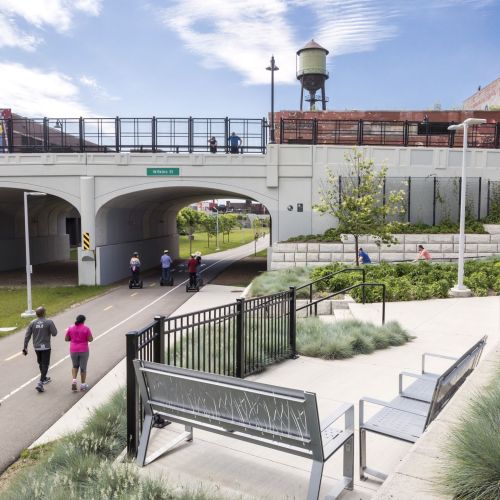 Walkers, bike riders, and segway scooter riders are sharing the non motorized roadway which is seen going under an overpass. To the right of the path are plazas with benches, cement walkways, grasses, and trees.