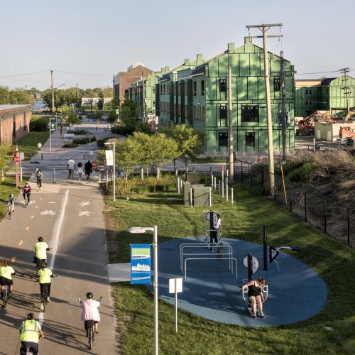 An aerial view of bikers on the non-motorized roadway. To the right, two people use outdoor exercise equipment in a designated area and in the background are buildings under construction. 