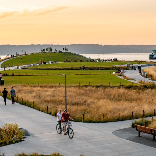 Pedestrians and bicyclists on wide paths surrounded by mown areas and prairie grasses with waterway in background.
