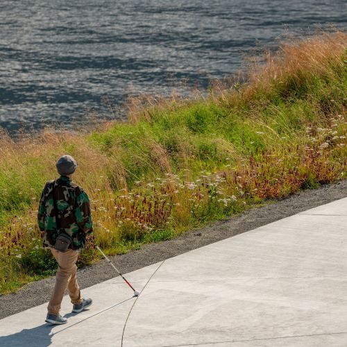 Person with white mobility cane walks along path with waterway in background.