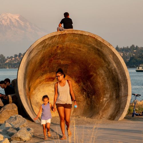 A child and adult walk in front of a large, round kettle-shaped structure with waterway in background