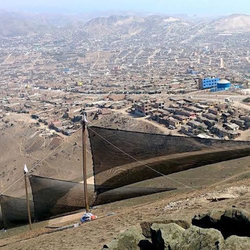 Large, triangular structures that appear to be made from mesh-like fabric are stretched taut with cables and resemble sails. Behind them are houses, with mountains in the background.