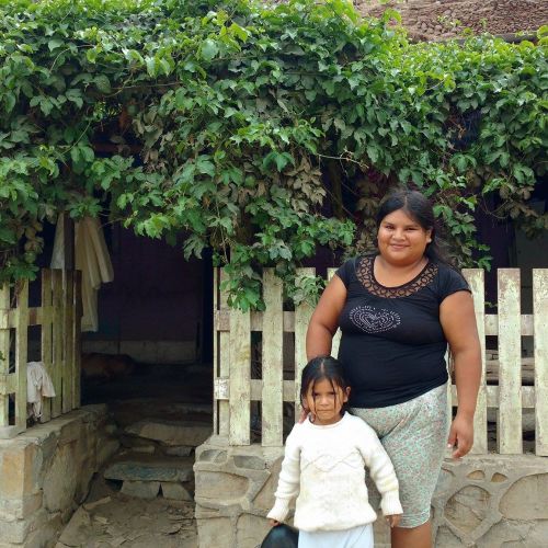 A young adult and child stand in front of a low stone wall with a white picket fence and lush greenery. Behind them are stone steps to an entryway.