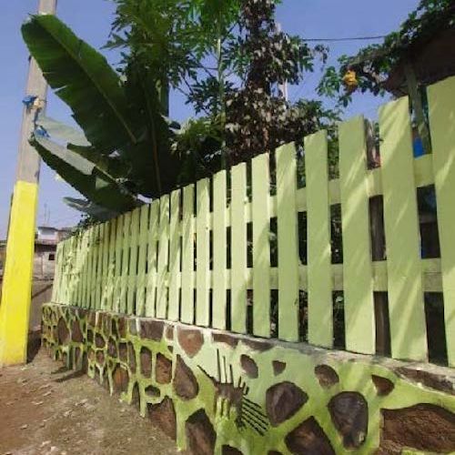 A picket fence painted pale yellow-green sits atop a stone wall with large plants behind it.