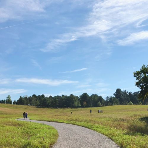 People walk on a wide, flat path with meadows on either side. There are trees in the background and an expanse of blue sky.