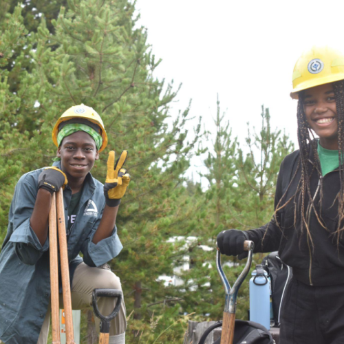 Two participants of color in the National Experiential Learning Program build a fence in front of a background of trees. They're smiling for the camera with tools in hand and wearing yellow hard hats.