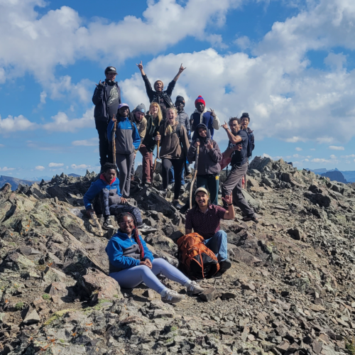 A group of participants pose for a picture on a rocky hill to commemorate one of Groundwork USA’s Yellowstone Experiential Learning trip groups in 2023. They're smiling broadly under a blue sky spotted with clouds. 