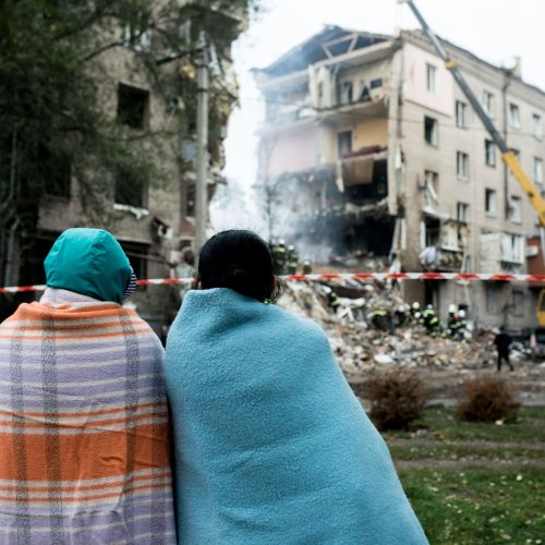 Two people with their backs to the camera have blankets draped across their backs and are  looking at bombed out buildings while standing on a patch of grass.