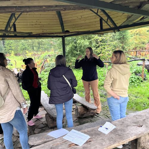 Group of adults gathered beneath an outdoor shelter in a garden with one adult leading the discussion.