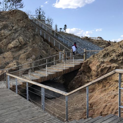 Two people walk up a wide wooden staircase. The staircase takes several turns as it goes between large rock outcrops.