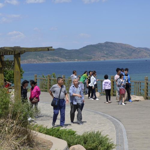 People walk along a wide path. There’s a body of water in the background with mountains behind it. 