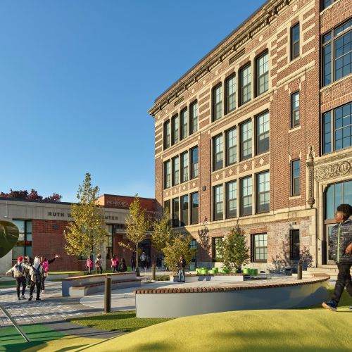 In front of the school, one student walks over the bright green berm while other students are welcomed to school by employees wearing bright pink vests. 