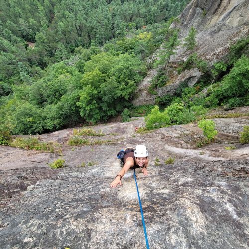 A young adult scales a steep rock face. The student is tethered by a rope and smiles at the camera with a sprawling forest below the cliff they are climbing.