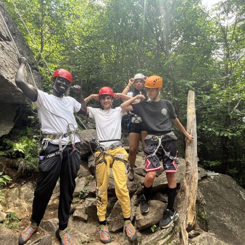 Four students of different races and genders flex their biceps. The students are in a forest next to large boulders, and are wearing helmets and climbing ropes and gear attached to them.