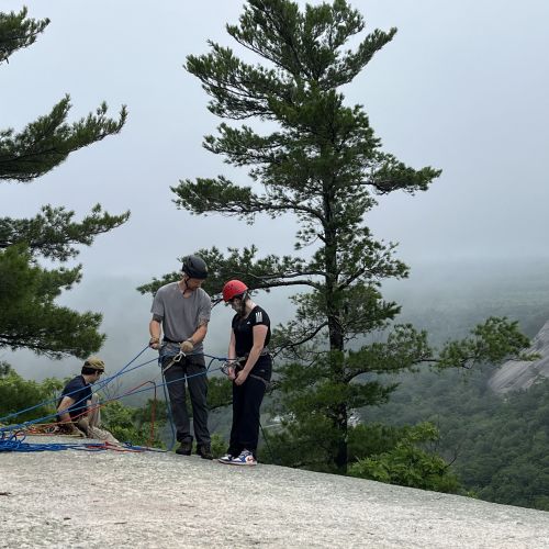 A group leader helps two students prepare their rock-climbing ropes. One sits on the edge of the rock while the other student prepares to repel. Large pines are adjacent to the students while a foggy mountain and forest are laid out in the distance.
