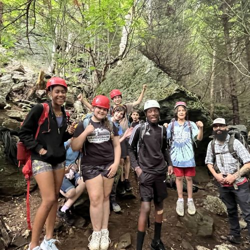 A bearded group leader poses with young adults of different ethnic backgrounds. They are wearing helmets and standing among boulders in a forested area.