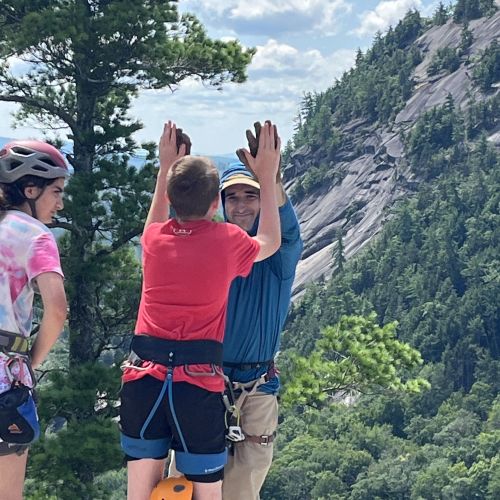 An adult male double-high-fives a young climber against the backdrop of a large, rocky mountainscape.