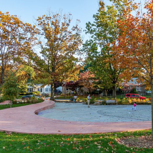 A salmon colored wall meanders through a park adjacent to lush green grass and open space. Several trees have leaves that are changing from green to orange. 