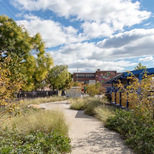A curved path is surrounded by grasses, perennials, and shrubs. On the right is a canopy structure with cars parked underneath.