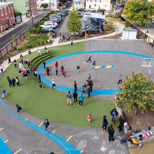An aerial view of the schoolyard showing people gathered along a curved path and berm around a basketball hoop where children are playing.