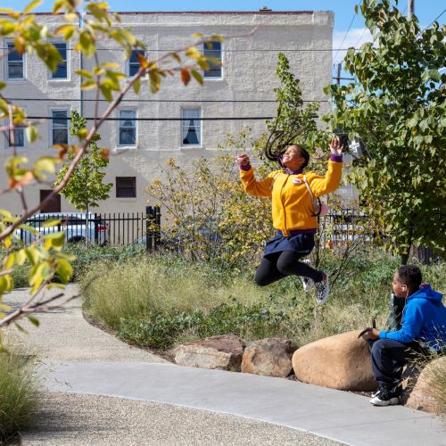 A smiling child jumps while another child sits on a boulder along a curved path surrounded by plants and trees.