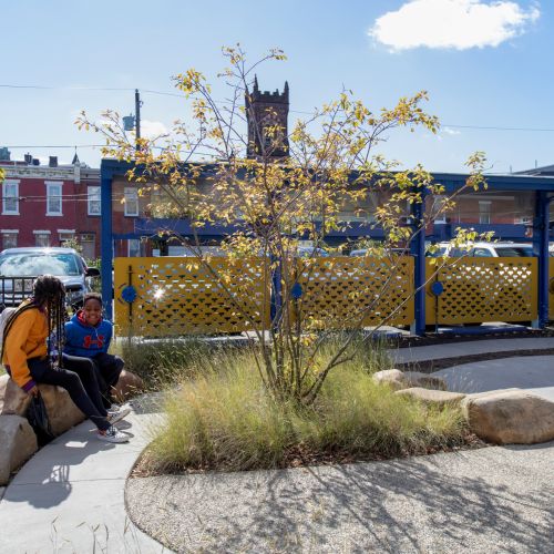 Children sit on boulders in front of decorative fence with parking area in background.