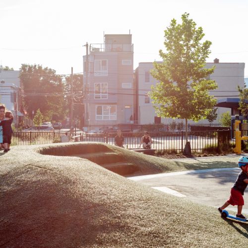 A woman kneels with a young child on the top of the berm while another child rides a scooter down the slope of the berm.