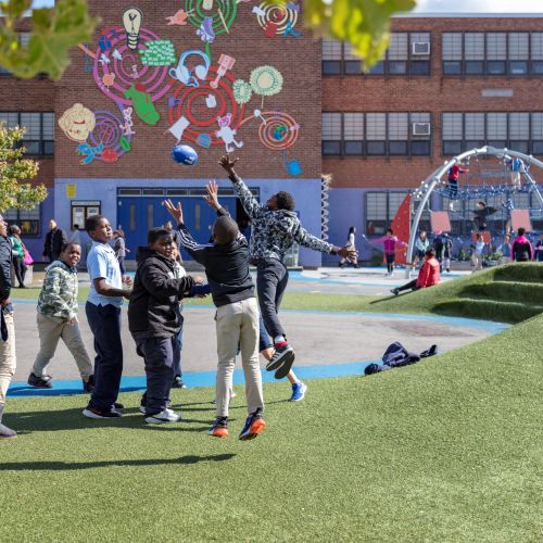 A group of youth play with a football while other children sit on the berm. Behind them, kids play on the climber dome. The school building in the background is painted with bright colors and artwork.