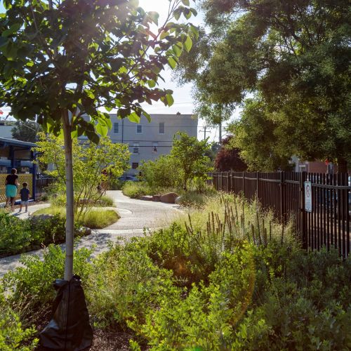 A child and adult walk along a curved path that is surrounded by trees and perennial gardens, with a fence on the right.