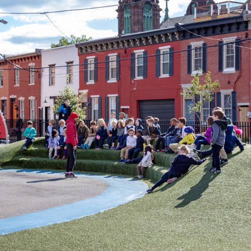 A classroom-sized group of young children sit on an artificial turf-covered berm facing toward an adult. 