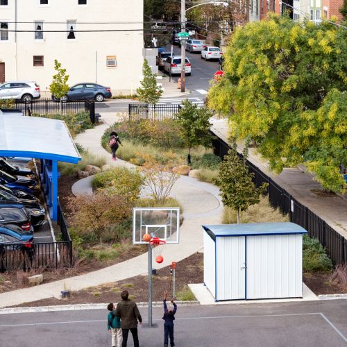 An adult and several children play basketball. In the background, a child walks along a curved path to a neighborhood street.