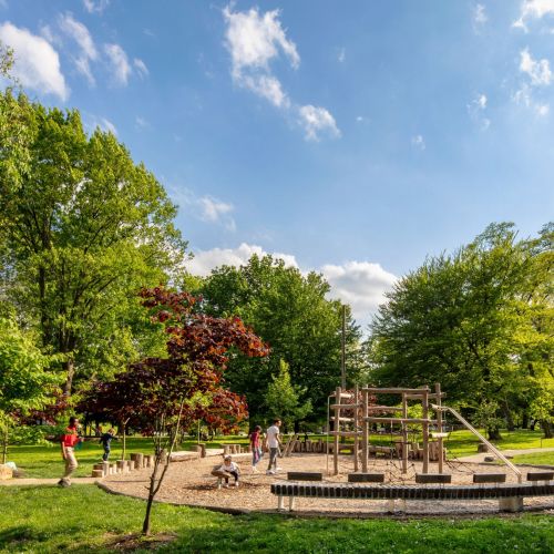 A circular perimeter made of natural materials surrounds a wooden play structure where children and adults are playing beneath a bright blue sky.