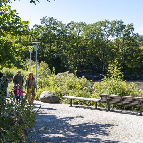 People walk along a gravel path with benches and a waterway in the background.