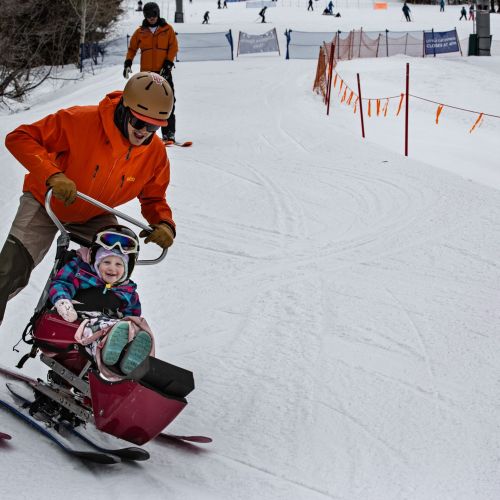 An adult in an orange jacket pushes a young participant down a gentle slope on a sit ski, which participants of all ages can enjoy.