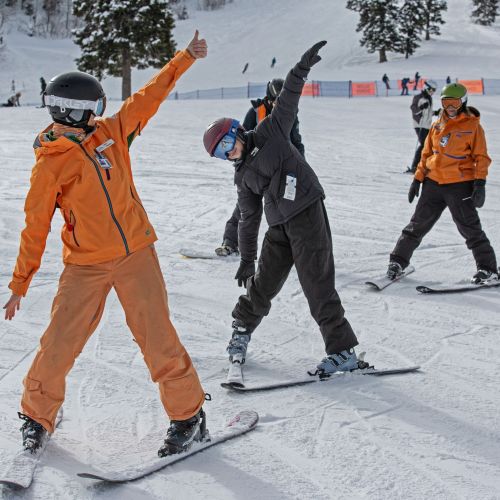 Two participants encourage each other with a thumbs-up as they prepare to go skiing with their peers. They are standing one behind the other with their arms spread out like airplanes. 