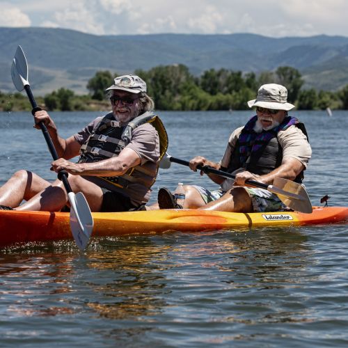 Two participants enjoy tandem kayaking in a lake, which is surrounded by lush trees and distant mountains.